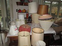 Photo showing multiple lamp shades of different sizes and styles on a wooden table, including beige, white, and red shades with wire frames.