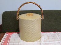 Front view of cylindrical ice bucket covered in cream grass cloth with bamboo handle and knob on lid, set on table.
