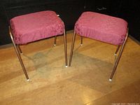 Two metal framed stools with pink patterned padded cushions, shown together from a side angle on a wooden floor in front of cabinets.