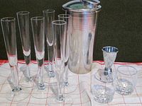 Six tall clear shot/champagne glasses, metal cocktail shaker, metal jigger, two Crown Royal shot glasses on a cloth surface.