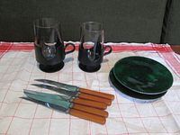Two black Corning cups, six knives with brown handles, and four green glass plates arranged on a red and white cloth