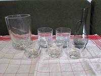 Seven clear glass items on a red and white checkered cloth including a small decanter with handle and spout, two wide and five different styled drinking glasses