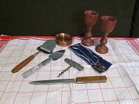Photo of all items on cloth including copper coasters, wooden goblets, letter opener in blue sheath, cake server, pie server, corkscrew, long knife.