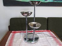Chrome modernist table lamp with three light cups shown on a wooden table with a cloth. Lamp has a rounded base and three vertical stems of varying heights topped with concave light cups.