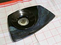 Front view of artisanal ikebana arranger with removable circular flower frog inserted into base tray showing glazed black and blue ceramic finish.