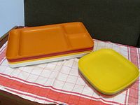Photo showing stack of 6 divided dinner trays in orange, yellow, and other colors, alongside 4 square plates in yellow and green vintage shades