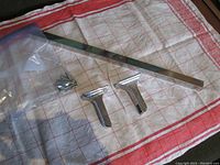 Photo showing two chrome finished wall holders, the straight metallic towel bar and mounting hardware (screws and anchors). Items laid on a cloth surface.