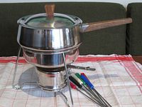 Photo of fully assembled fondue set showing stainless steel pot with teak wood handle and lid, placed on metal burner stand with warming bowl underneath, along with colorful fondue forks set.