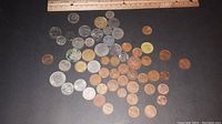 Photo showing an assortment of around 50 coins spread on a dark surface with a ruler for scale above them. Coins vary in size and color from silver-toned, copper, and gold-tone metals.