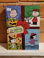 Front cover of the deluxe Peanuts holiday collection box with colorful illustrations of Charlie Brown, Snoopy, and Lucy in holiday scenes.