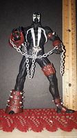 Front view of 7-8 inch Spawn action figure standing upright, showing black and white suit, brown spiked gauntlets and boots, with silver chain held in hands.