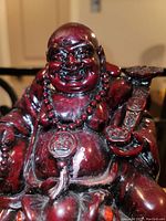 Front view of red laughing Buddha statue showing facial details, beaded necklace, and ruyi scepter.