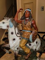 Figurine of an indigenous man wearing tan clothing and a blue headband, riding a white spotted horse.