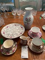 View of china cake plate, multiple floral teacups and saucers, small pitcher, and tall floral vase on wooden table
