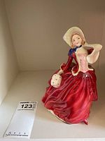Porcelain figurine of a woman in a long red dress and bonnet holding a basket, shown with a scale sticker.