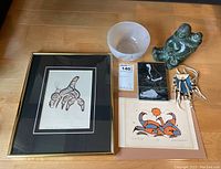 Overview photo showing framed Norval Morrisseau print, signed art by Norval Morrisseau, stone sculpture with crack, black and white stone etching, frosted bowl, and beaded medicine pouch.