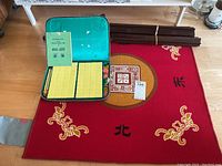 Overview photo showing the green carrying case with arranged yellow-backed mahjong tiles, red table cover with Chinese characters and decorative motifs, and four dark brown wooden tile stands stacked on floor.