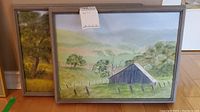 Two framed rural landscape oil paintings leaning against wall on floor. Both depict barns with trees and hills.