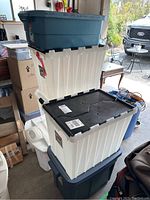 Stack of assorted plastic storage bins including large white translucent bins with black lids and large dark blue bins with solid lids