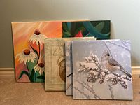 Photo showing six canvas artworks stacked against a wall, partially overlapping, revealing variety of designs including flowers, bird, abstract and coffee cup