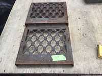 Two rectangular metal air registers with diamond lattice patterns placed on a surface. Visible rust and wear indicate age and use. Dimensions approximately 12-1/4 x 13-3/4 inches.