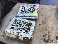Photo of two vintage metal air registers with white paint, rust, and ornate cut-out designs on a wooden surface