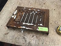 Cast iron decorative air register vent cover with rust and peeling paint on wood surface.