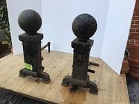 Two cast iron fireplace andirons with spherical finials seen from front on wooden surface with white background
