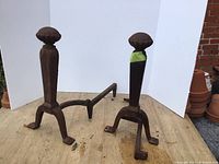 Photo of two antique cast iron fireplace andirons on wooden board, showing detailed finials and legs.