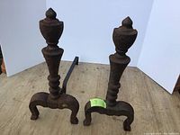 Pair of antique cast iron fireplace andirons standing upright on a wooden surface with white panels behind, showing decorative urn finials, turned columns, and curved legs.