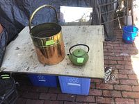 Copper bucket and green iron teapot on a worktable with blue recycling bins underneath.