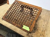 Photo of the rusted metal floor register vent cover lying on a wooden surface with a green lot number tag.