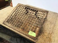 Photo of a metal floor register vent cover with visible rust and dirt, placed on a wooden surface