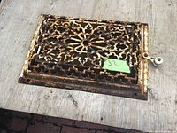 Top view of a rectangular decorative cast iron vent cover with ornate lattice work and adjustment lever hub resting on wooden surface.