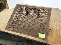 Cast iron floor register showing detailed geometric and floral cutout pattern with signs of surface rust laid on wooden surface.