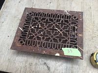 Photo showing rectangular cast iron floor register with geometric decorative pattern, rusted surface, and minor paint splatters on a wooden floor.