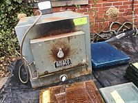 Front view of the square metal kiln showing the front door, brand name 'Amaco Electric Kiln', temperature control knob, power cord, and stacked glass plates around it.