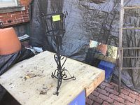 Metal plant stand with ornate scroll design sitting on a wooden table outdoors near a brick wall and tarp.