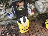 Yellow Karcher pressure washer with power cord wrapped around base placed on brick surface, surrounded by paint cans and stone items.