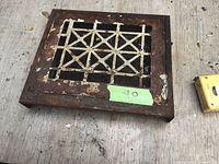 Rectangular metal register vent cover with cross and parallel bar pattern showing rust and paint chipping.