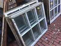 Two wooden window sashes with six glass panes each, resting against a pile of old wood and metal objects, showing several layers of old paint and wear.