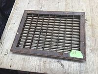 Steel vent register placed on a wooden surface showing its full rectangular grid and frame with visible wear.