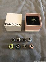 Nine glass charm beads displayed on fabric with a Pandora box next to them. Colors include green, pink, purple, blue, yellow, and metallic hues. One bead is inside a pink velvet jewelry box with Pandora white box beside it.