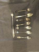 Top view of 8 sterling silver spoons and 1 serving fork, displayed on dark cloth, showing various handle designs and enamel medallions.