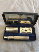 Open compact case holding the three disassembled pieces of the Gillette safety razor: handle, inner guard, outer guard. Case has a mirror and blue velvet lining.