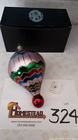 Glass hot air balloon Christmas ornament with colorful zigzag stripes next to black box and tag, displayed on printed paper with lot number 324.