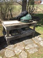 Full view of Big Green Egg grill in wood table stand on stone patio, black cover draped over dome