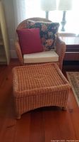 Wicker chair with two throw cushions and a beige seat cushion, plus matching woven footstool in front on wooden floor with furniture background.