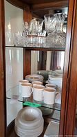 Photo shows glassware including faceted crystal decanter and various styles of glasses - wine, rock, and gold-rimmed glasses on a cabinet shelf.