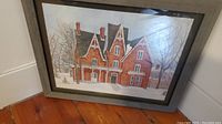 Framed print of a large red brick country home in winter with snow and leafless trees, shown leaning against white wall on wood floor.